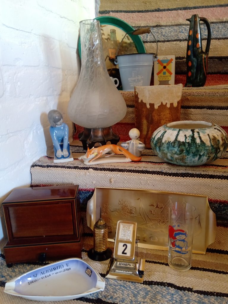 Különféle retro és vintage használati tárgyak gyűjteménye egy lépcsőn elhelyezve, köztük porcelán figurák, egy asztali naptár, üvegedények és egy dekoratív tálca.
